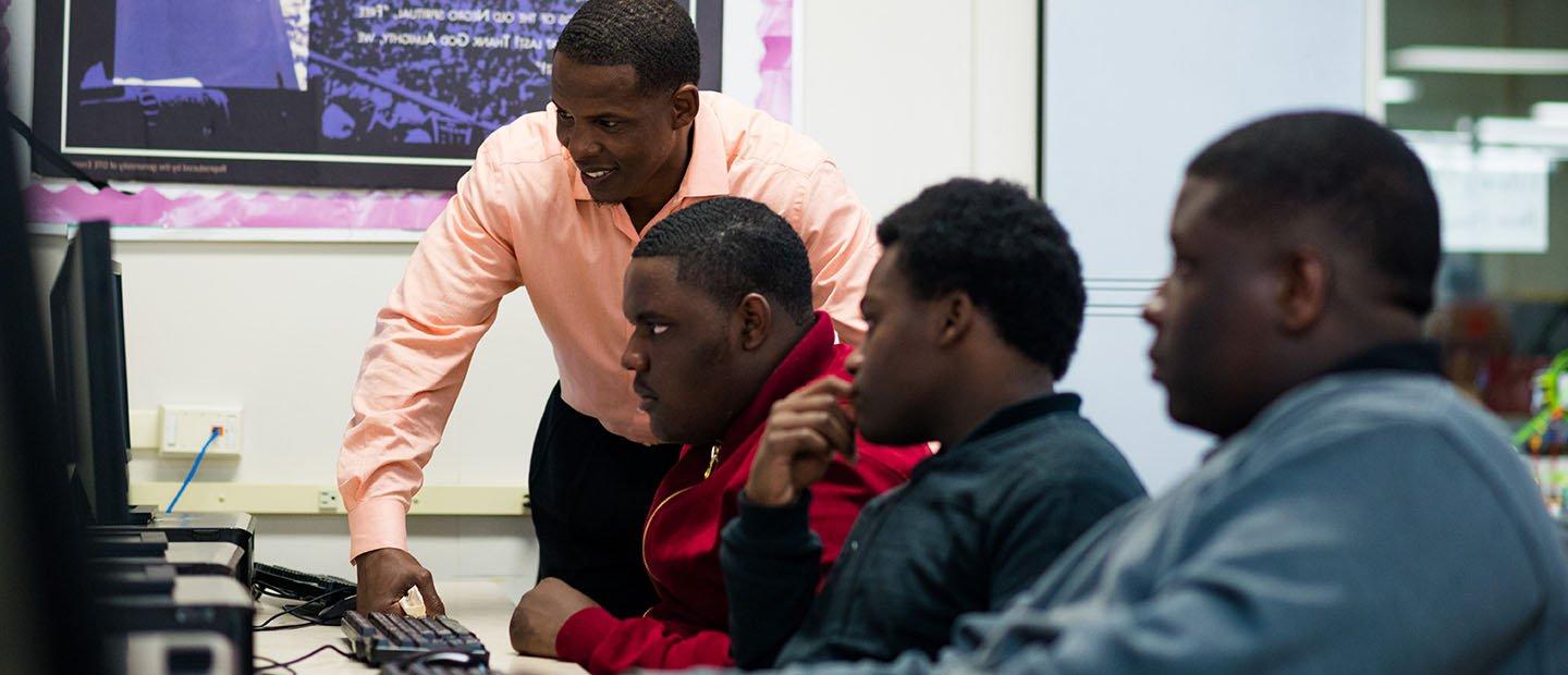 A man standing over a row of three young men working at computers.